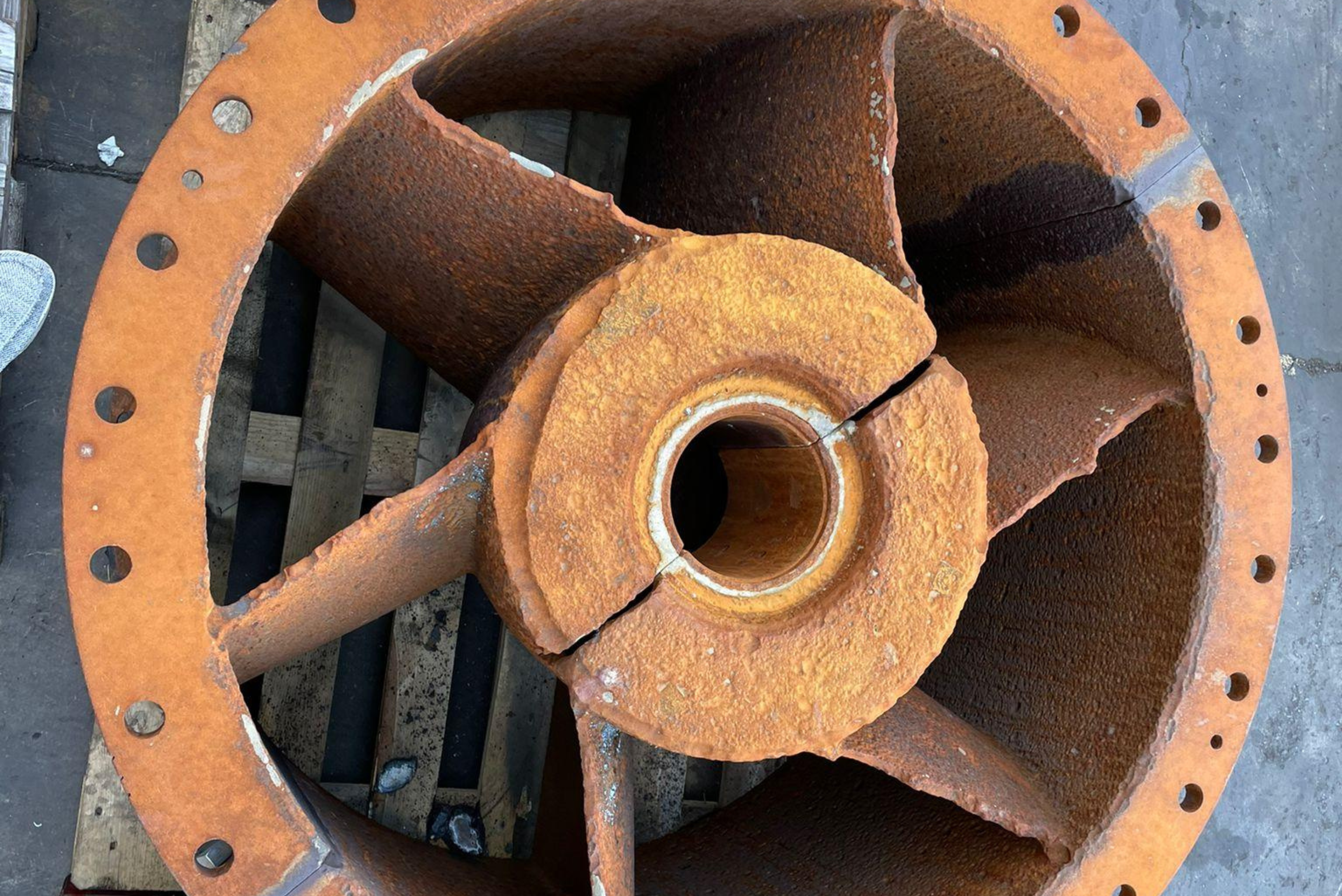 Vue de face d'un composant industriel circulaire fortement rouillé, doté de pales intérieures et d'un moyeu central perforé, posé sur des palettes en bois.