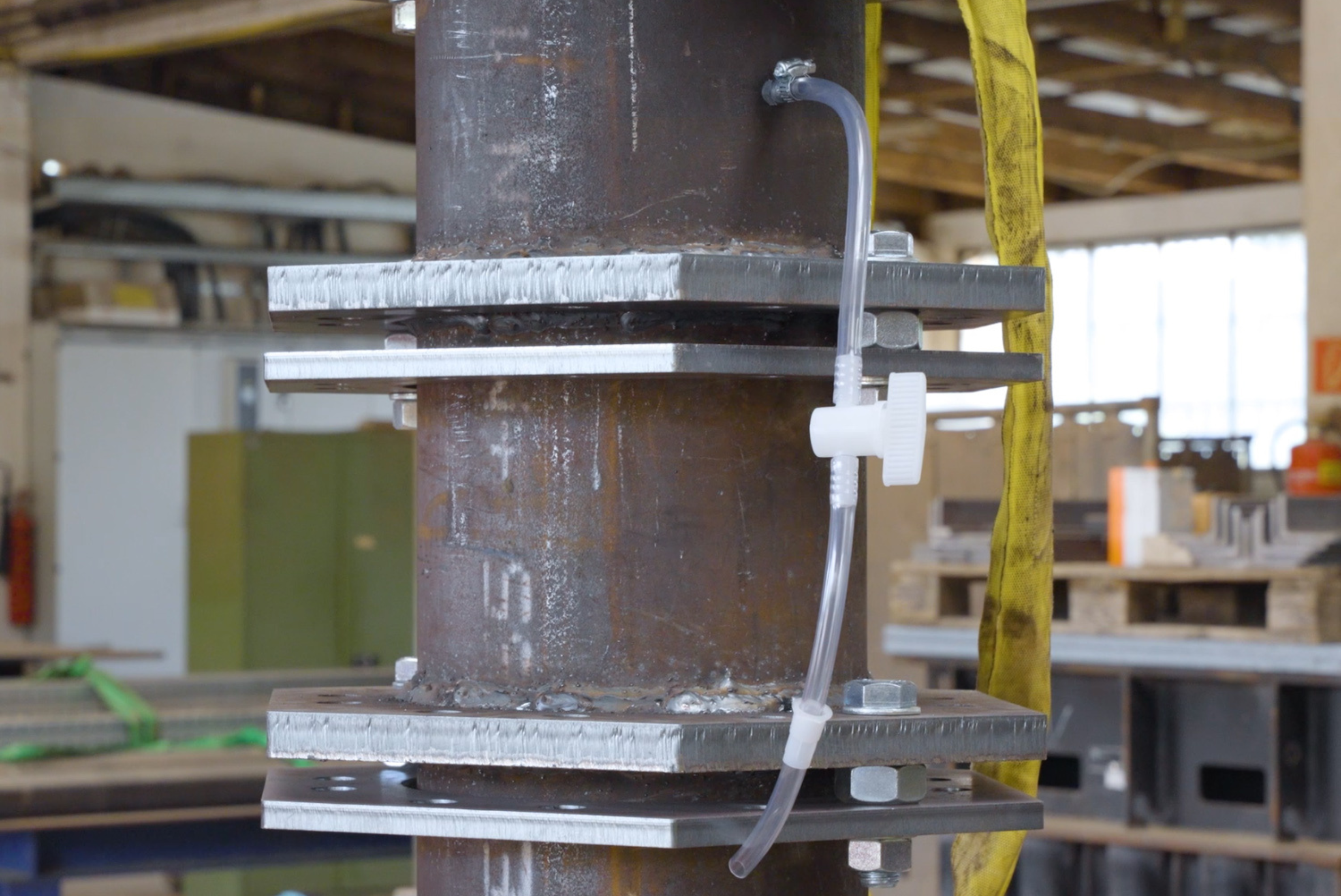 Une colonne métallique industrielle verticale reliée par des plaques à brides épaisses et boulonnées, avec un petit tuyau transparent équipé d'une vanne blanche fixé le long de la structure.