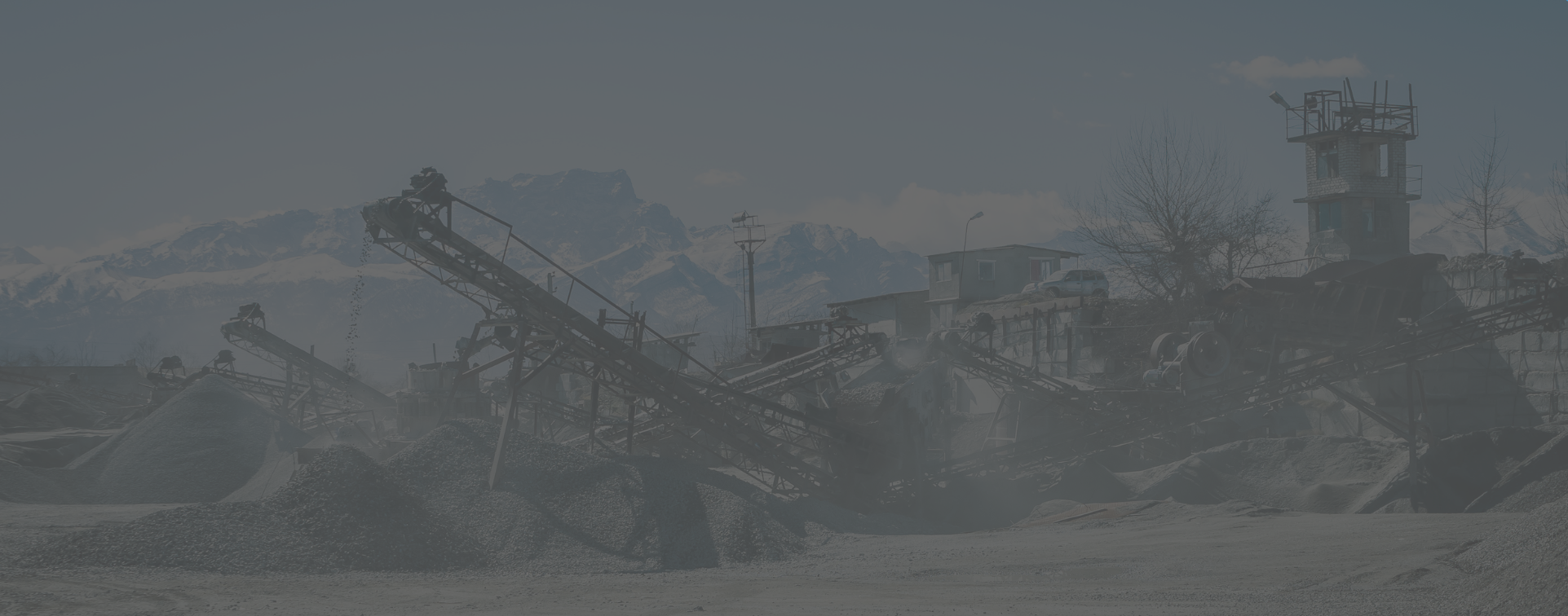 Vue d'ensemble d'une grande installation industrielle de concassage et de convoyage avec des bandes transporteuses de matériaux en vrac, des tas de gravier et des structures de bâtiments sur fond de montagnes.