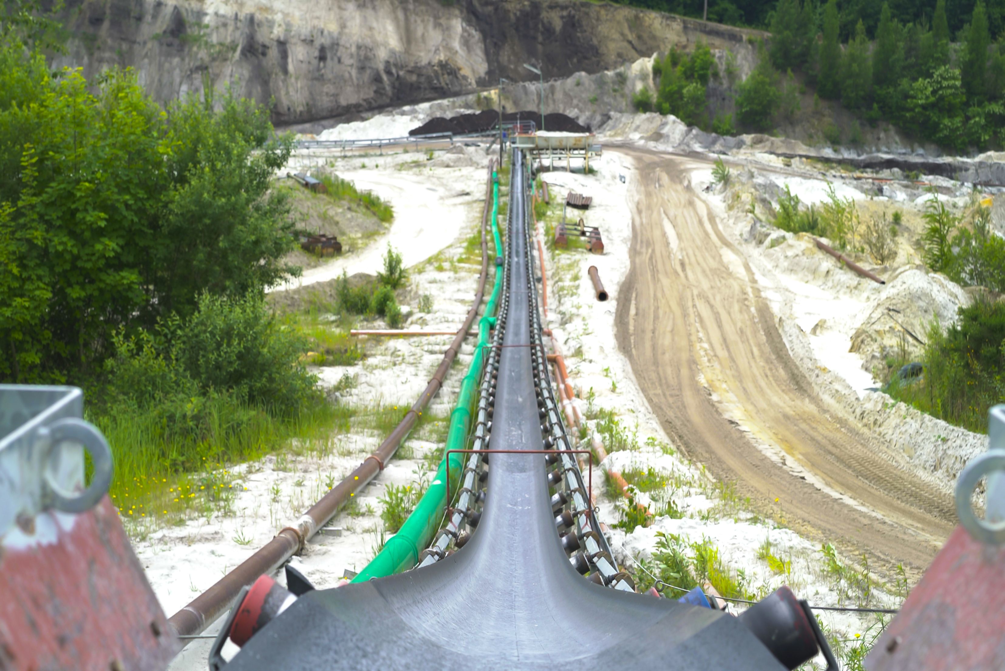 Une longue bande transporteuse industrielle s'étendant à travers un vaste paysage minier ou de carrière, bordée de verdure et de chemins de terre.