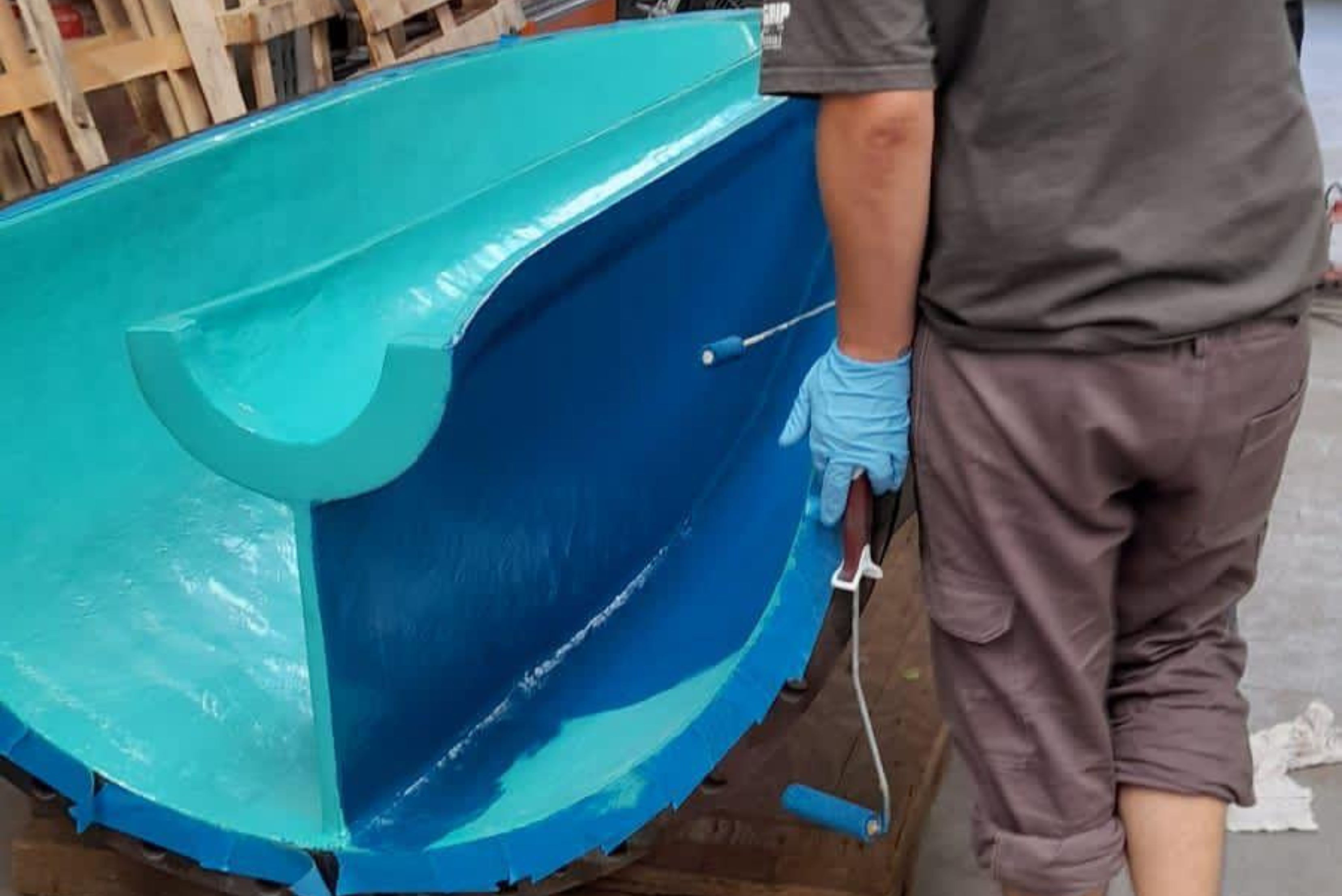 Un ouvrier portant des gants de protection applique un revêtement bleu sur une grande structure métallique semi-cylindrique à l'aide d'un petit rouleau.