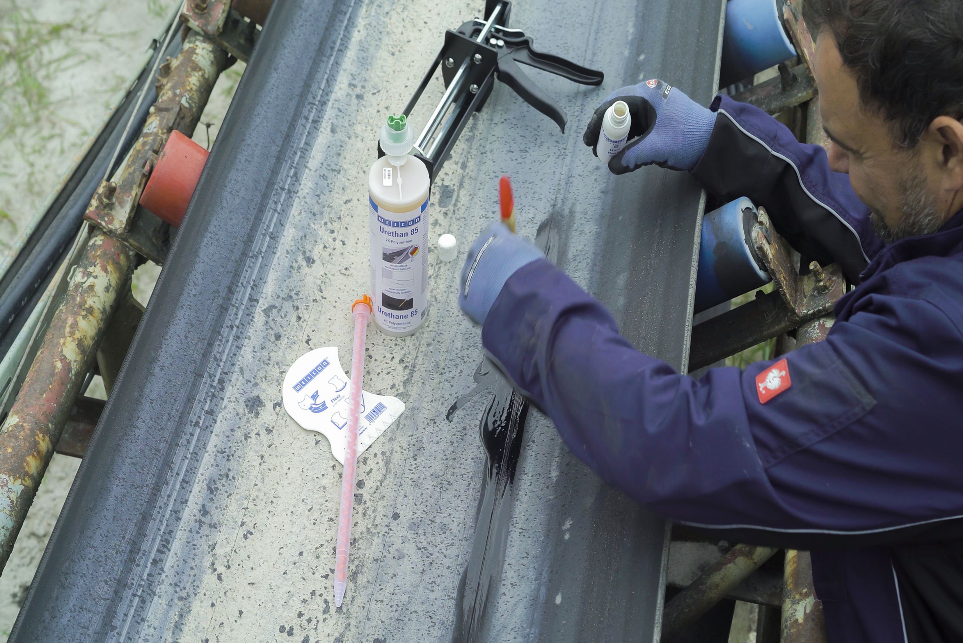 Un technicien portant des gants applique de l'adhésif ou du mastic à l'aide d'un petit pinceau sur une bande transporteuse en caoutchouc, entouré de flacons de produits chimiques et d'un pistolet à calfeutrer.