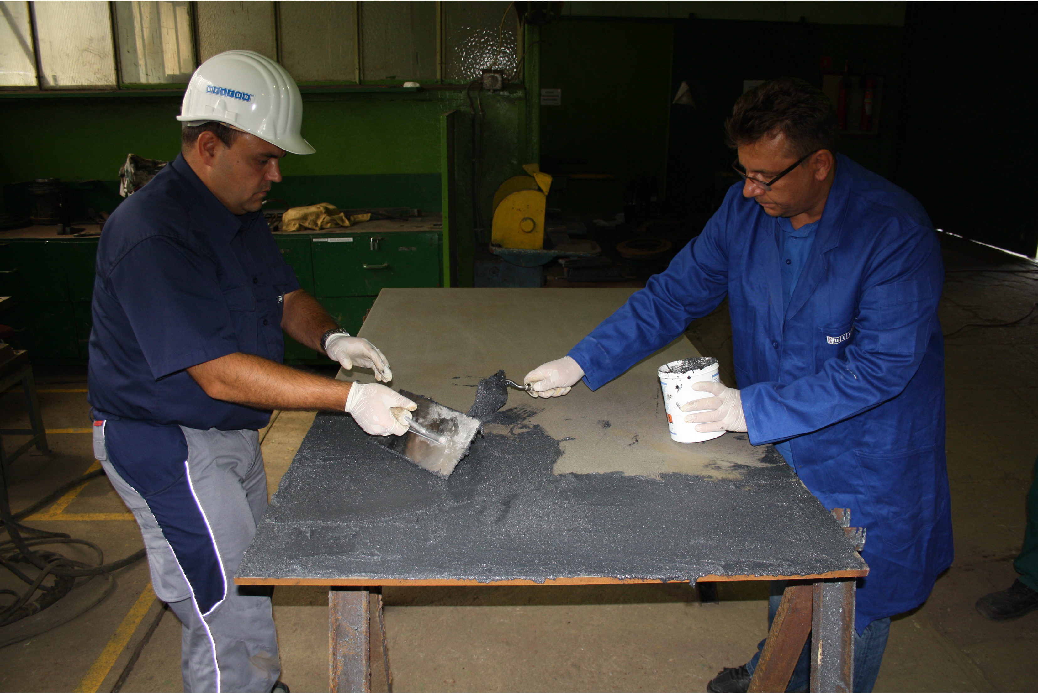 Deux techniciens en vêtements de travail et gants de protection appliquent une masse de résine sombre sur une grande plaque métallique posée sur des tréteaux dans un atelier.