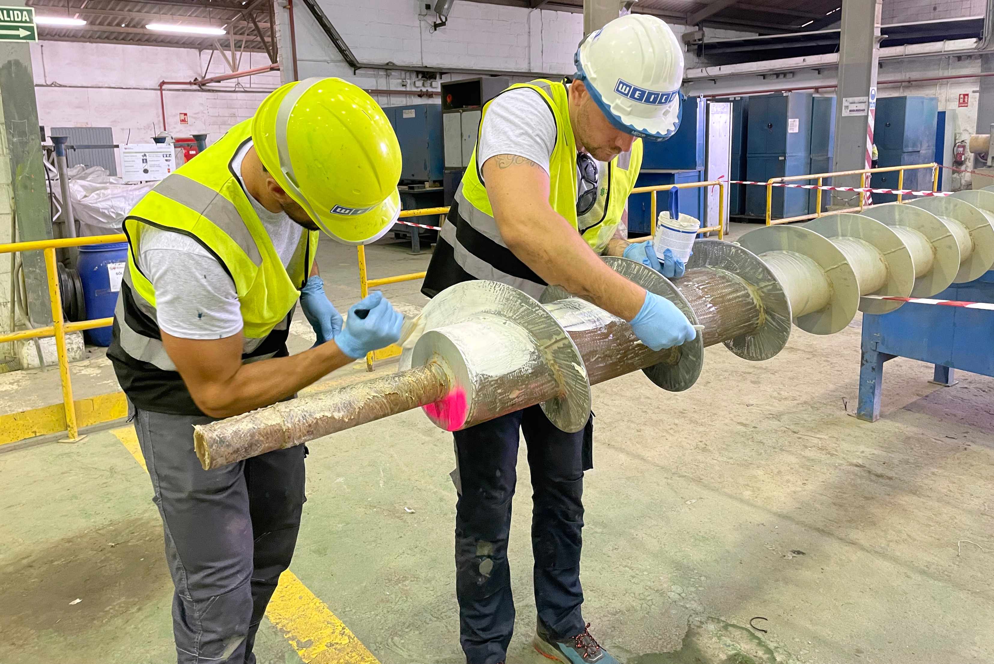 Deux ouvriers en gilets de sécurité et casques appliquent un revêtement sur une grande vis sans fin industrielle dans un atelier.