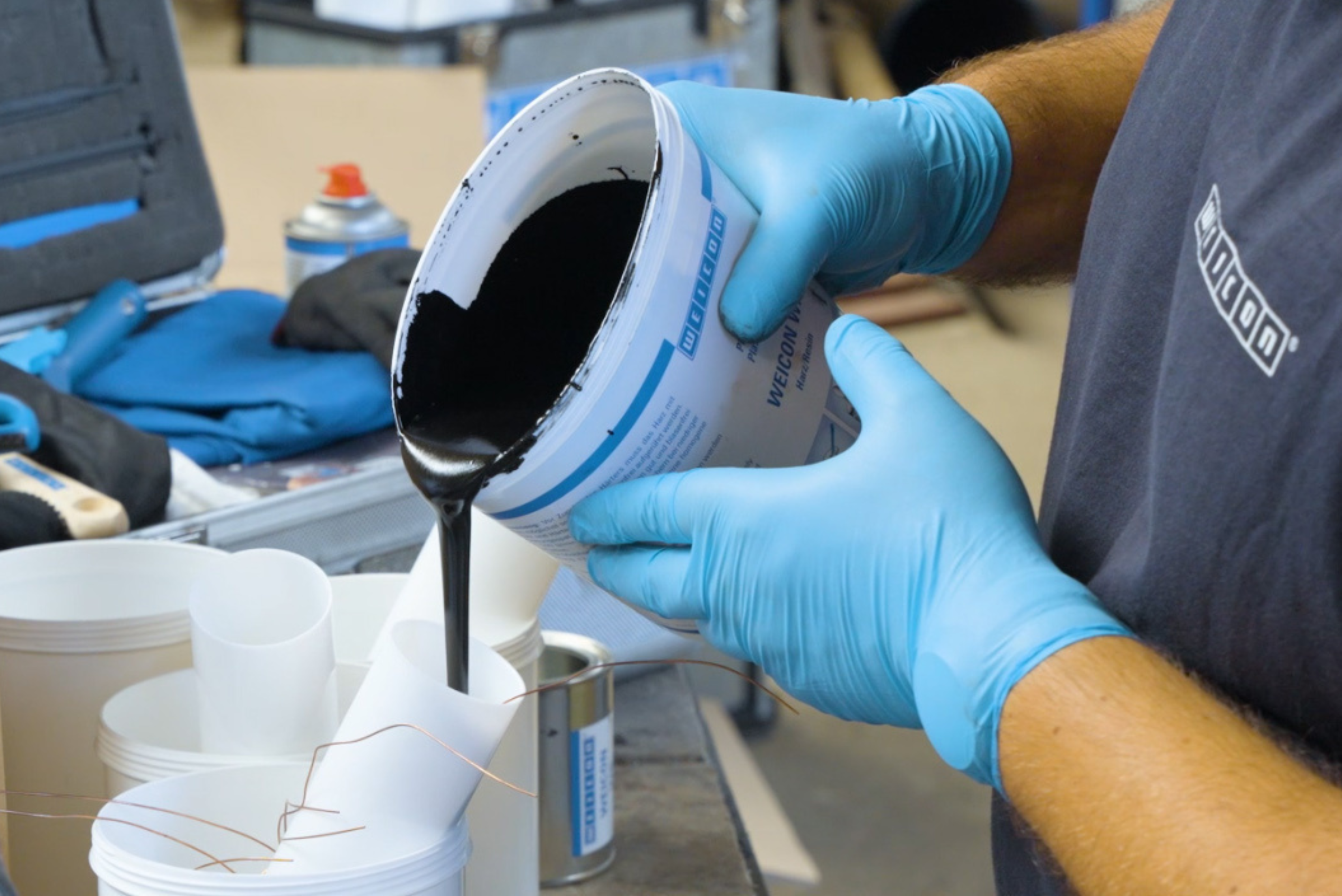 Une personne portant des gants de protection bleus verse une substance sombre et visqueuse d'un seau portant l'inscription WEICON dans un petit gobelet blanc, tandis que d'autres récipients sont disposés au premier plan.