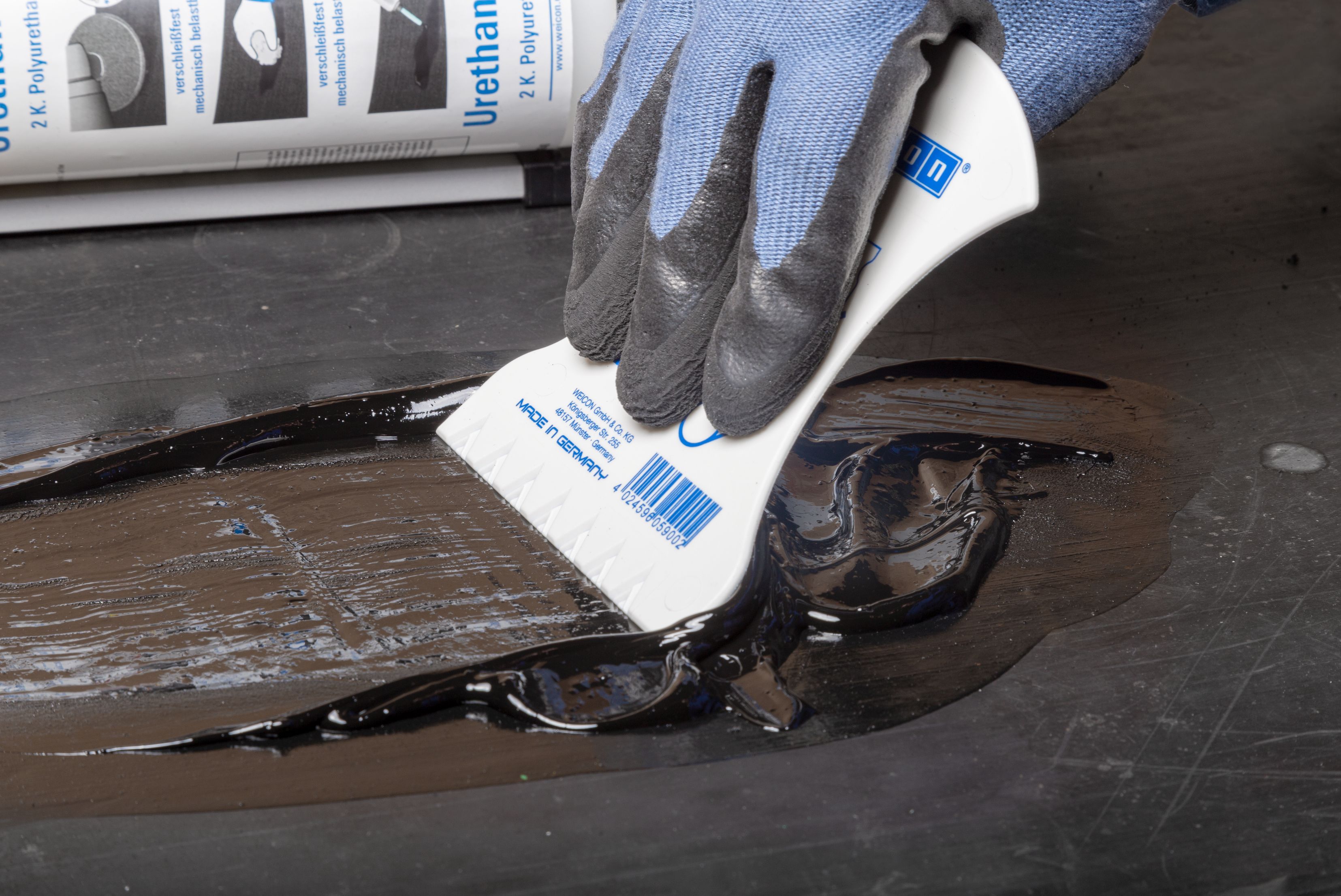 Une main gantée utilise une spatule en plastique blanche pour étaler et lisser un produit de réparation noir et visqueux sur une surface sombre, avec une cartouche de produit visible en arrière-plan.