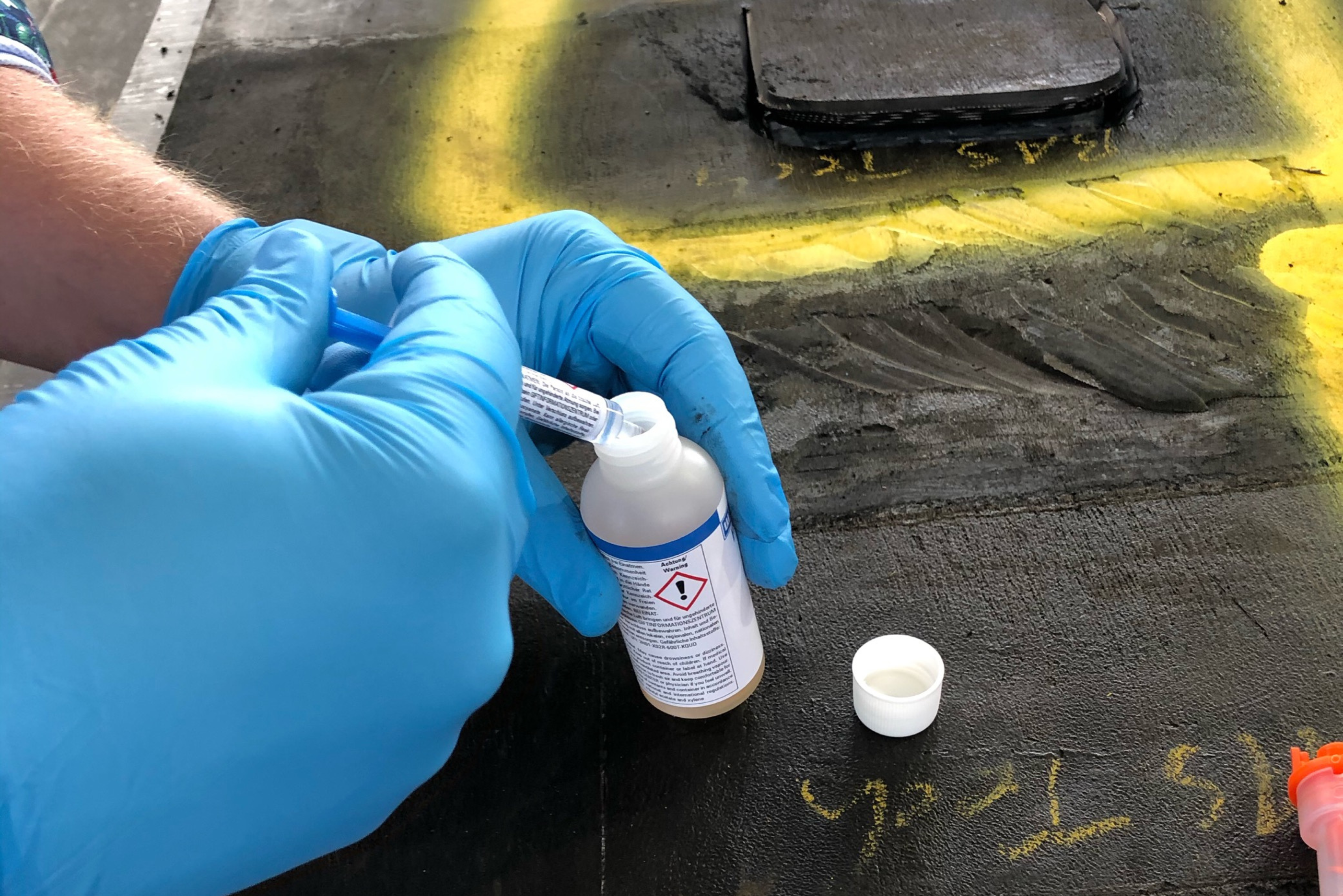 Une personne portant des gants bleus utilise une petite seringue ou un applicateur pour ajouter un liquide provenant d'une petite fiole étiquetée sur une surface sombre avec des marquages jaunes.