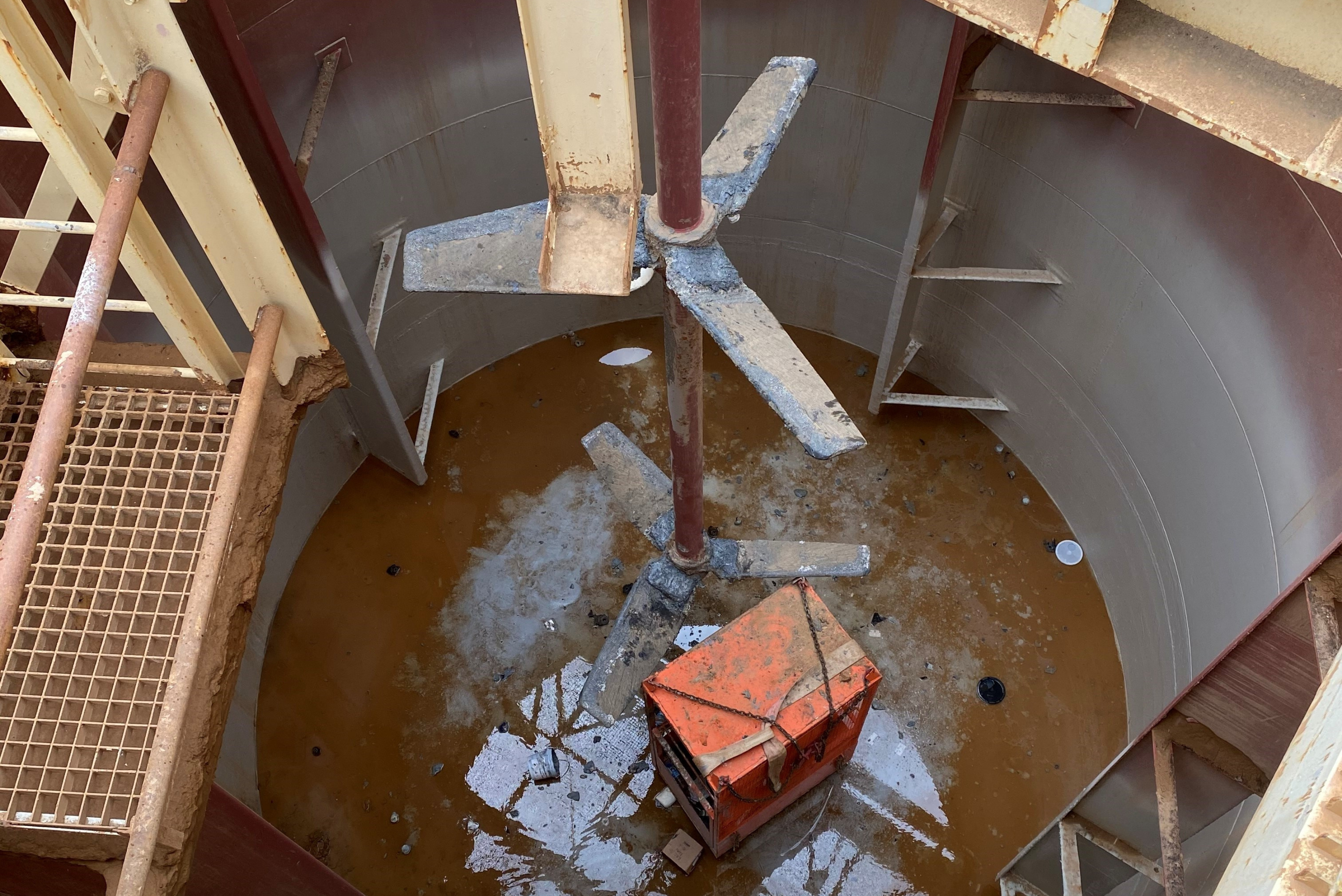 Une vue en plongée d'un grand réservoir ou d'une cuve industrielle cylindrique contenant un liquide brun, avec un agitateur vertical à plusieurs pales en son centre et une caisse ou une palette orange posée au fond