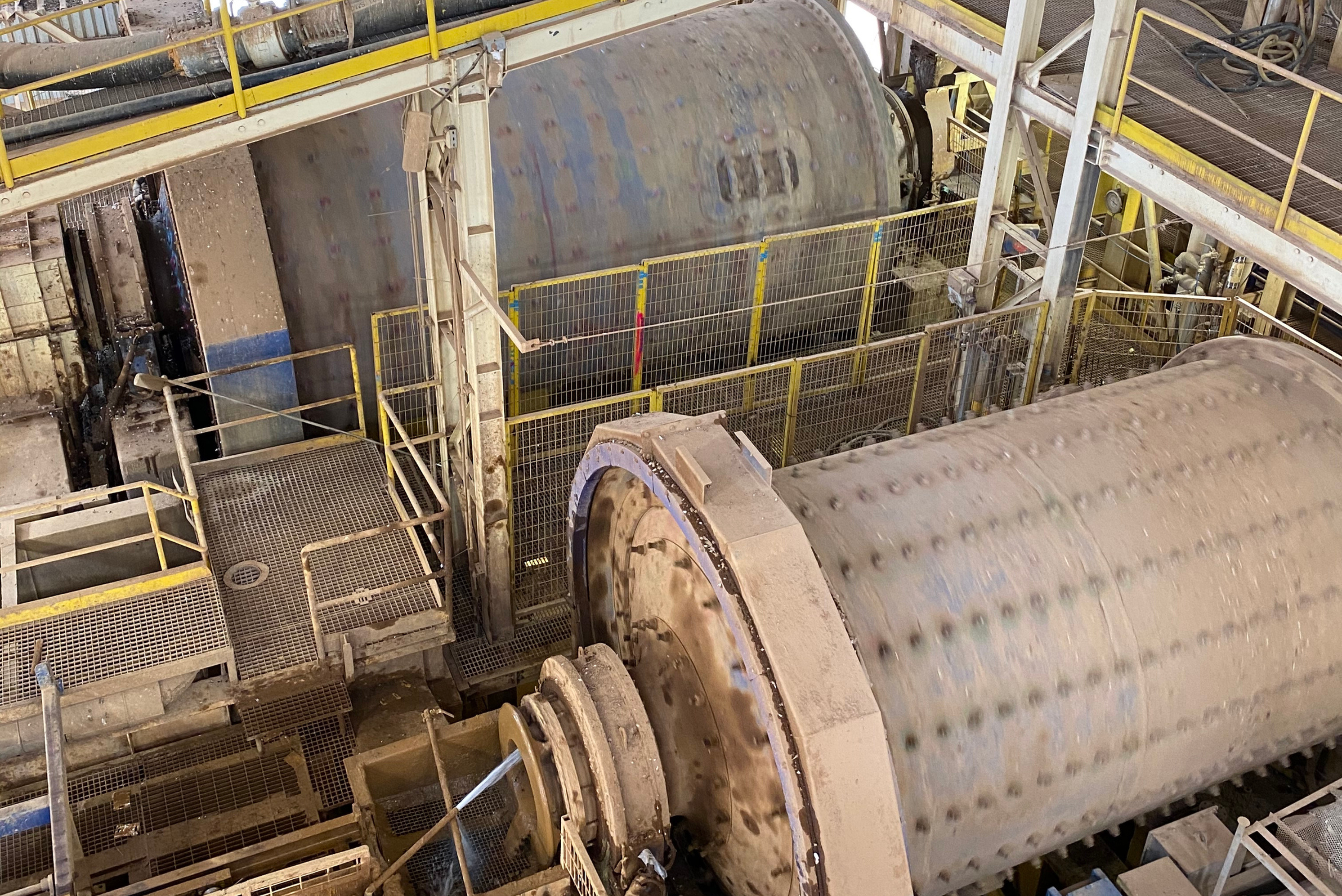 Une vue en plongée d'une grande usine industrielle avec deux grands broyeurs à boulets cylindriques ou tambours de traitement des minerais, entourés de passerelles métalliques et de barrières de sécurité jaunes.