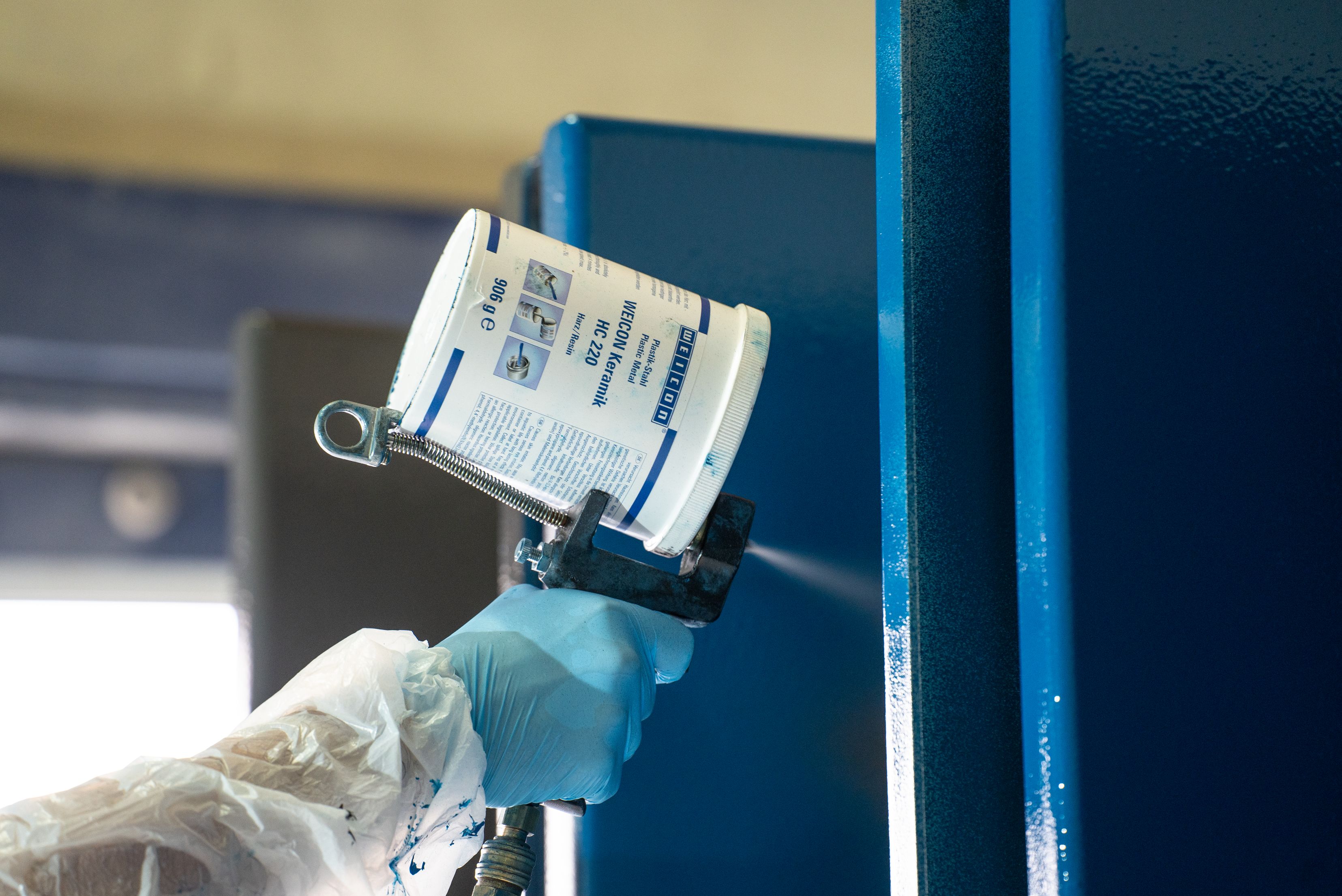Gros plan sur une personne portant des gants de protection et une blouse blanche, utilisant un pistolet de pulvérisation doté d'un godet fixé pour appliquer de la peinture sur une surface métallique bleue.