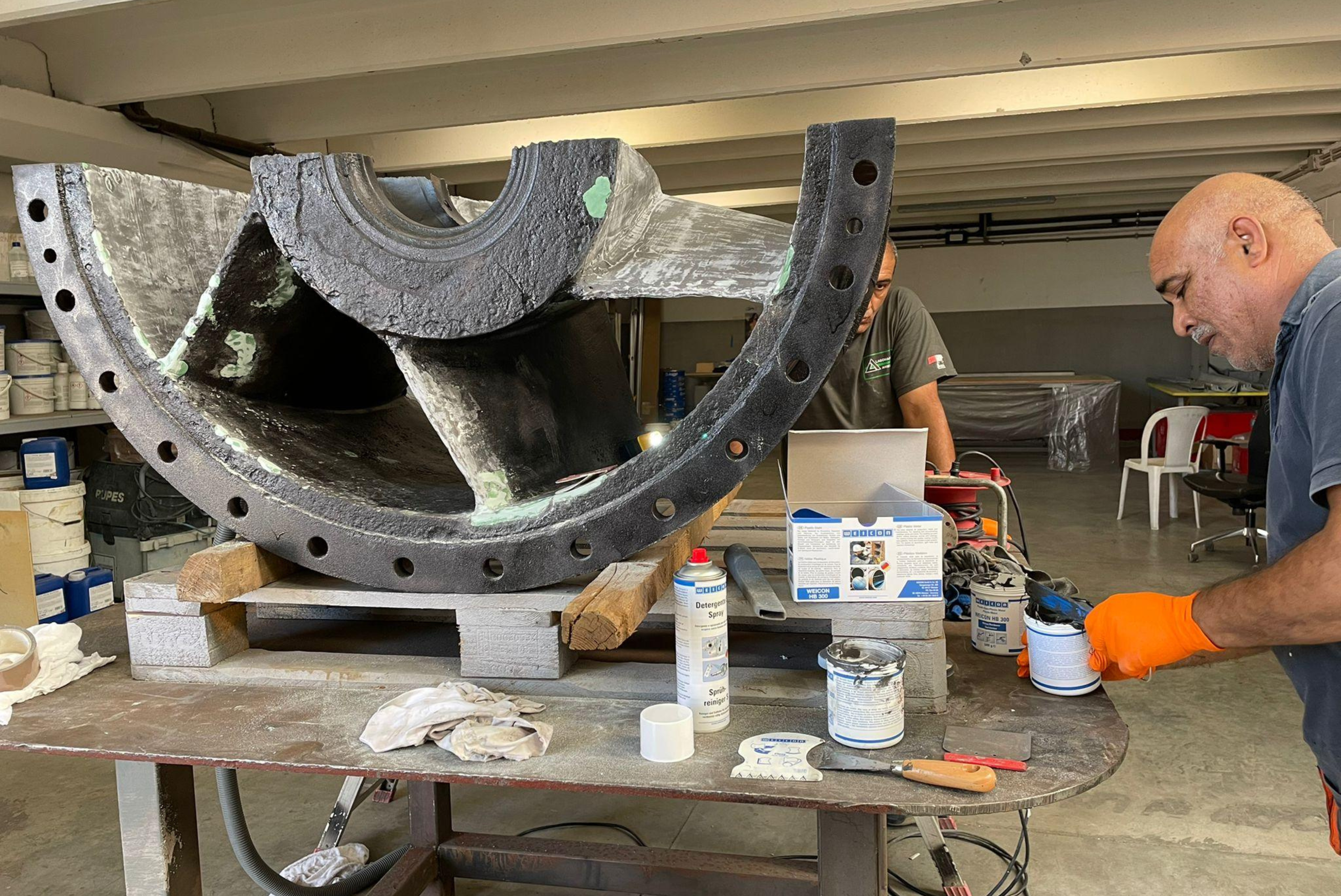 Vue d'ensemble de travailleurs dans un atelier préparant et mélangeant des produits de revêtement à côté d'une grande pièce industrielle semi-circulaire posée sur des palettes.