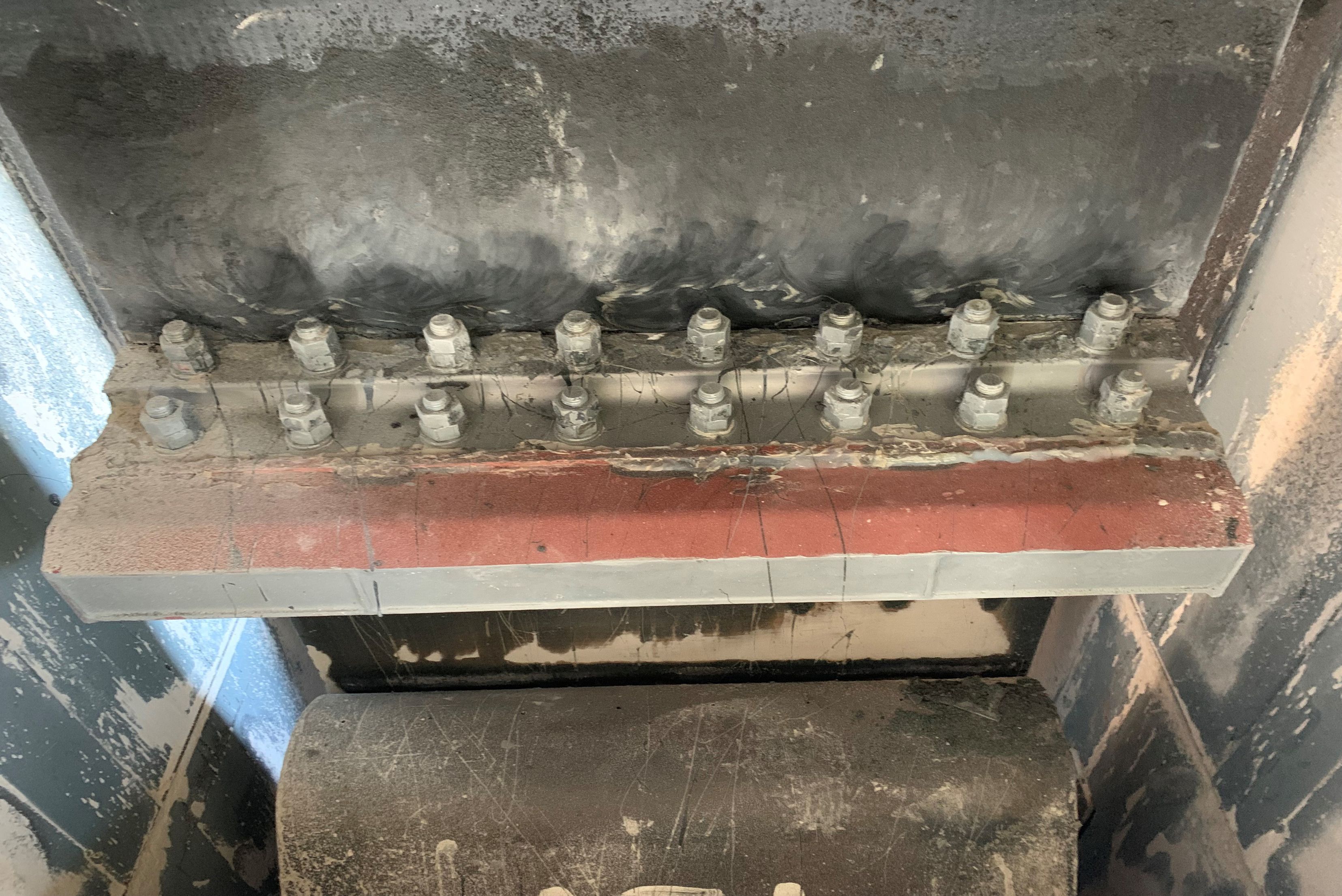 Vue rapprochée d'une structure industrielle métallique montrant deux rangées horizontales de boulons et d'écrous fixant des plaques de métal robustes, avec une surface sombre et poussiéreuse en arrière-plan et un cylindre ou rouleau métallique en bas.