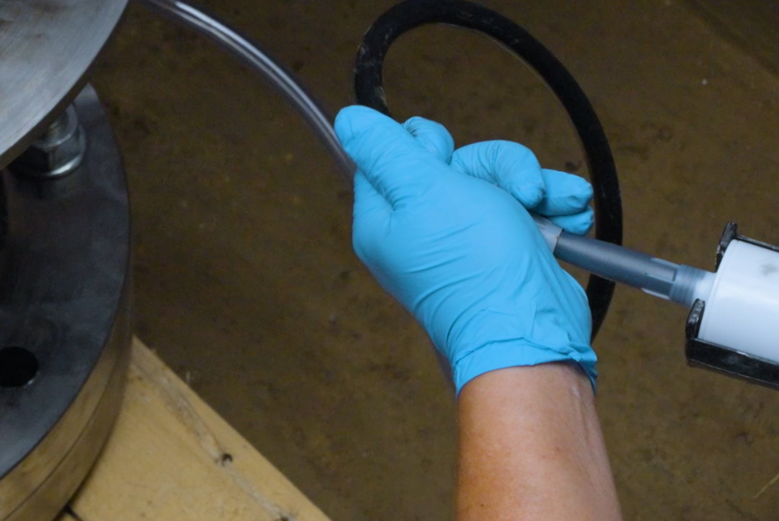 Une personne portant des gants de protection bleus tient une cartouche ou un applicateur de produit d'étanchéité relié à un tube fin, tout en manipulant un joint torique noir à proximité d'une bride métallique.