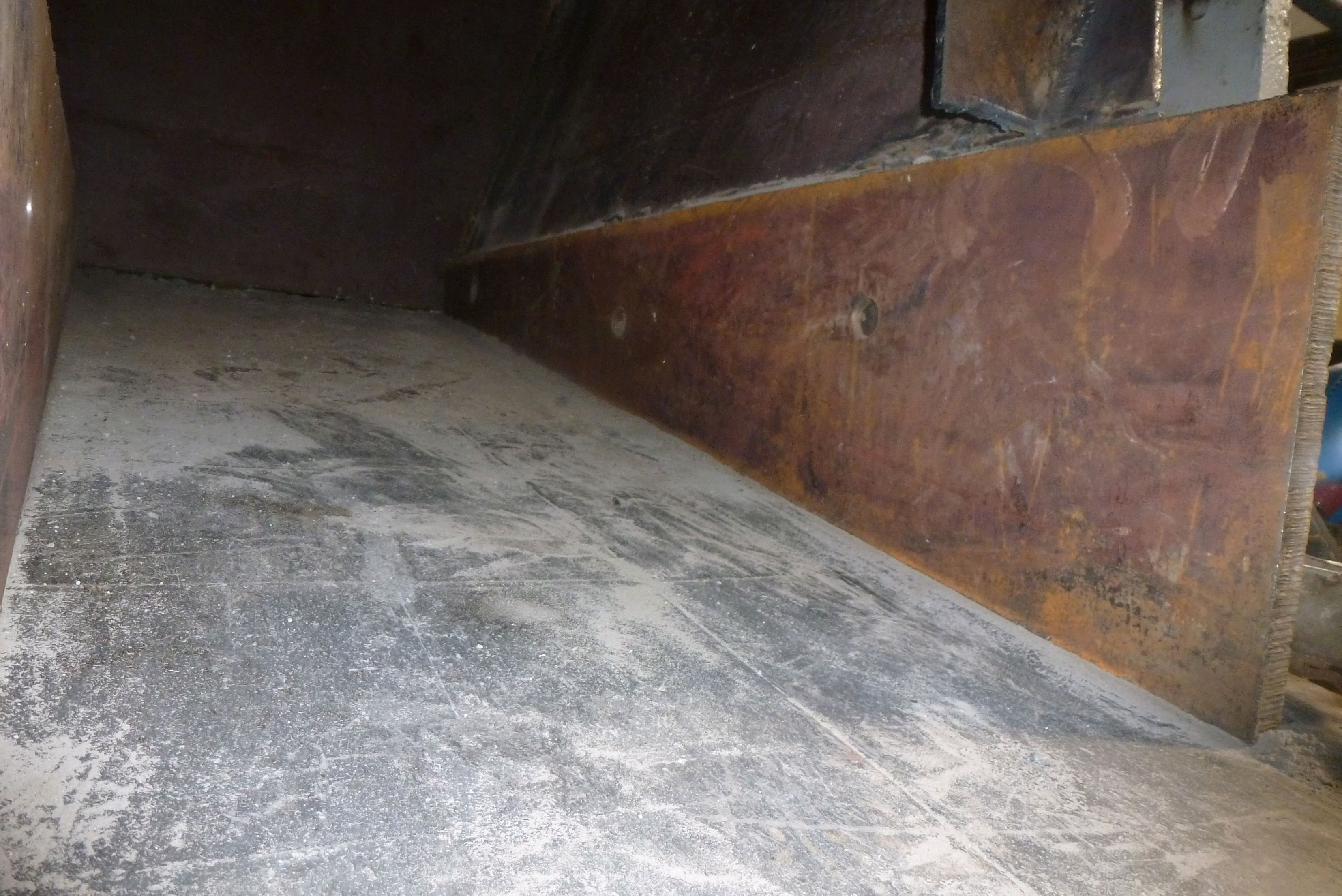 Vue rapprochée de l'intérieur d'une trémie de déchiqueteuse ou d'un convoyeur, montrant une plaque latérale en acier fortement rouillée fixée au-dessus d'une surface en acier grise et usée, couverte de poussière.