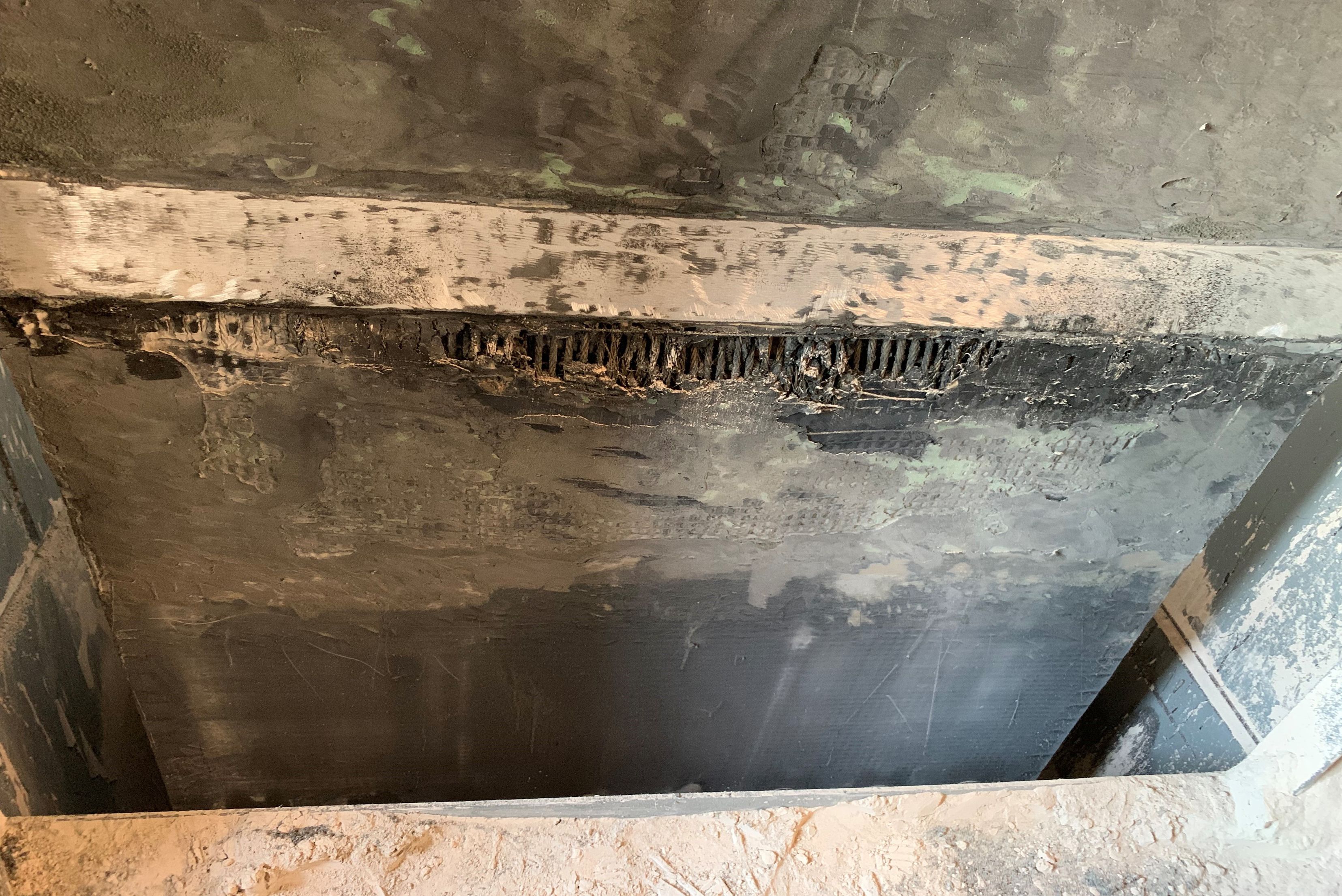 Vue rapprochée en contre-plongée de l'intérieur d'une structure industrielle ou d'une goulotte, montrant des surfaces métalliques sombres et poussiéreuses avec des traces d'usure et des résidus sombres le long d'un joint ou d'une ouverture.