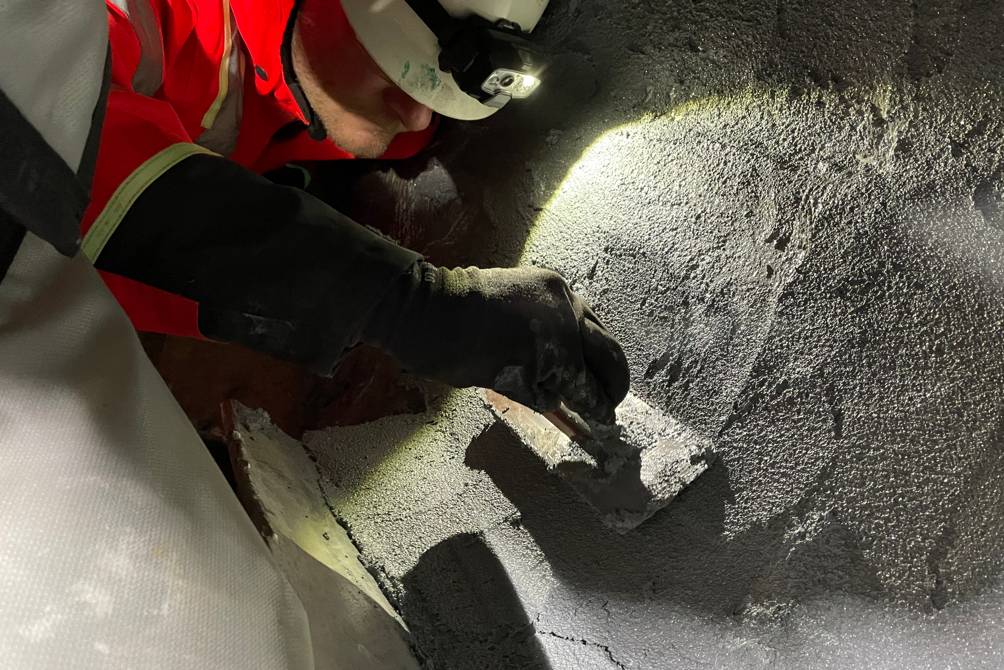 Gros plan sur un ouvrier avec un casque et une lampe frontale qui applique du mastic ou un revêtement protecteur sur une surface métallique granuleuse à l'aide d'une truelle.