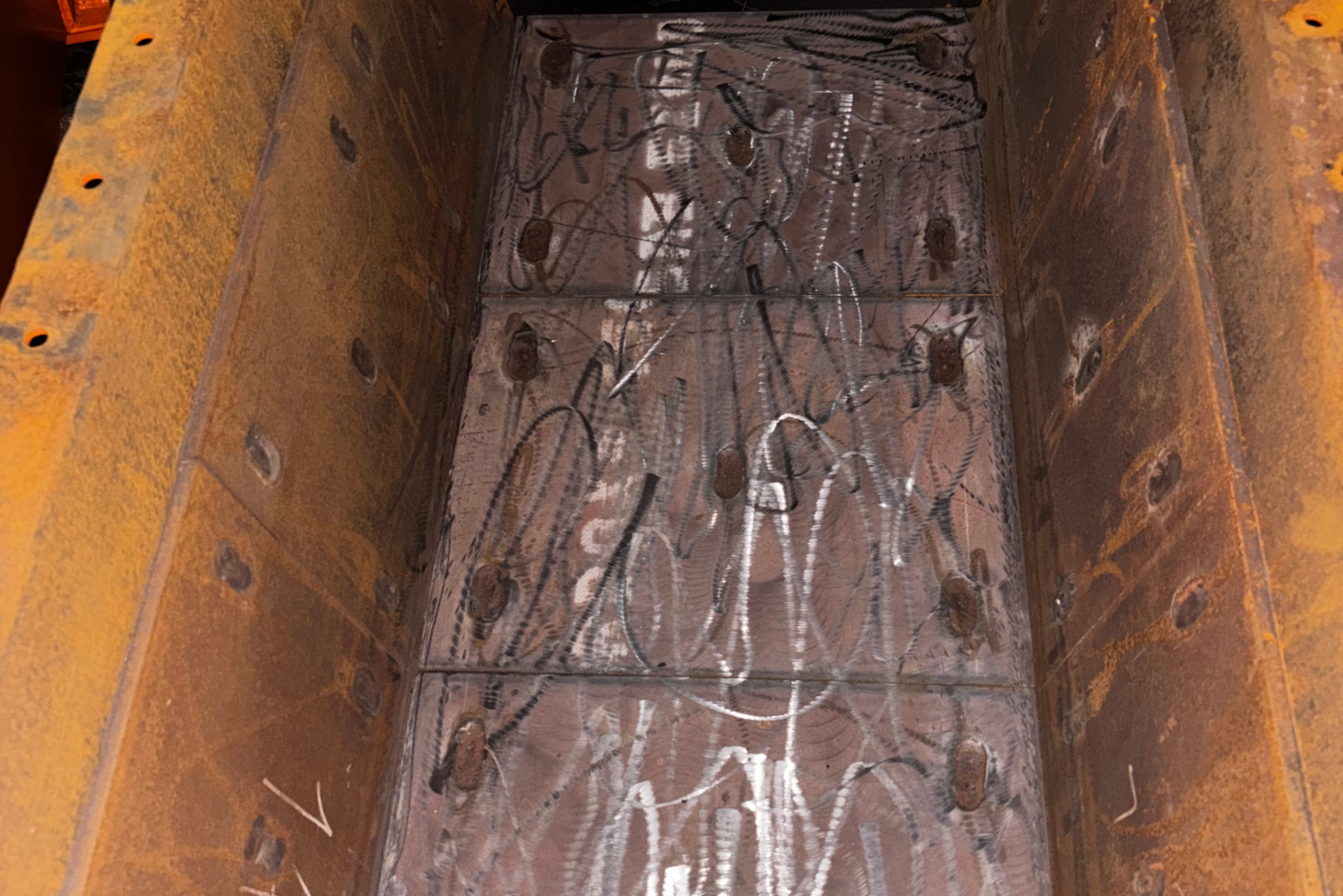 Une vue en plongée d'une goulotte ou d'un canal industriel en métal rouillé, avec des surfaces internes usinées ou traitées montrant des traces de meulage et des rangées de fixations par boulons.