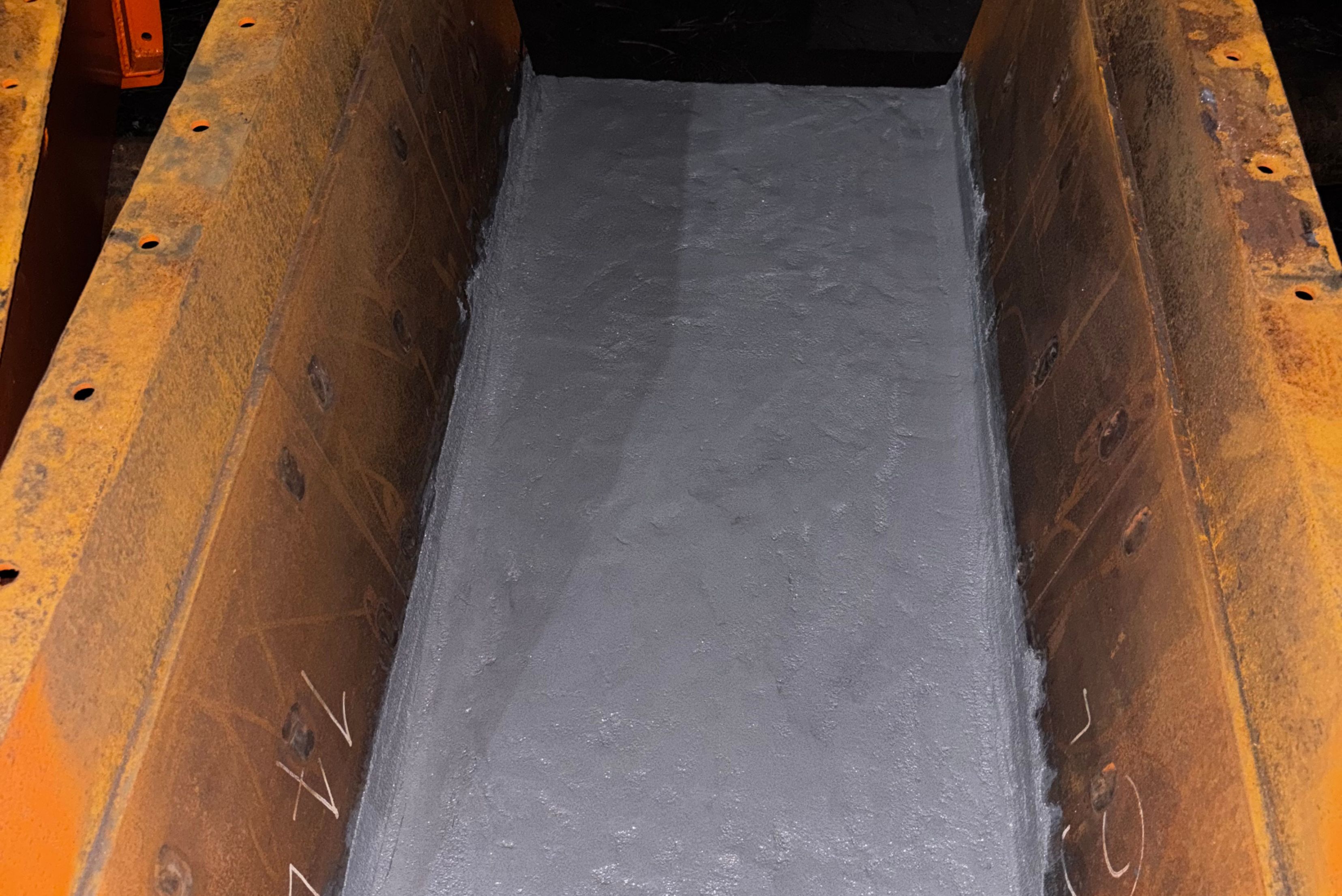 Une vue en plongée d'un canal ou d'une goulotte industrielle en métal, dont le fond intérieur est entièrement recouvert d'une couche de revêtement de protection ou de résine gris clair et lisse, encadrée par des parois latérales brun rouille.