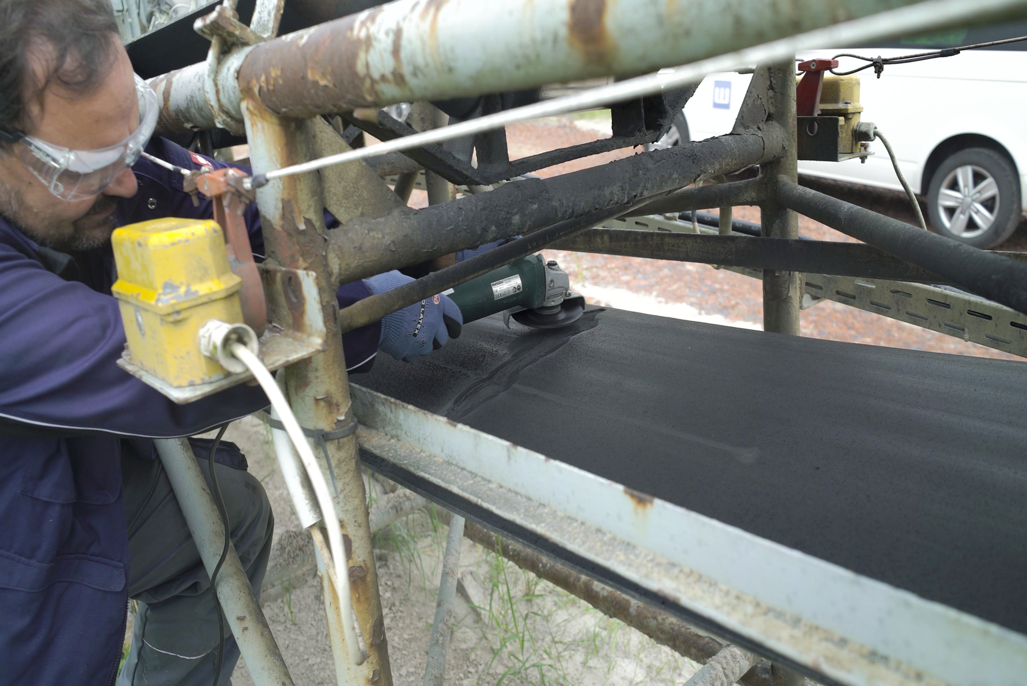 Un technicien portant des lunettes de protection et des gants utilise une meuleuse d'angle pour travailler sur une grande bande transporteuse industrielle en caoutchouc noir.