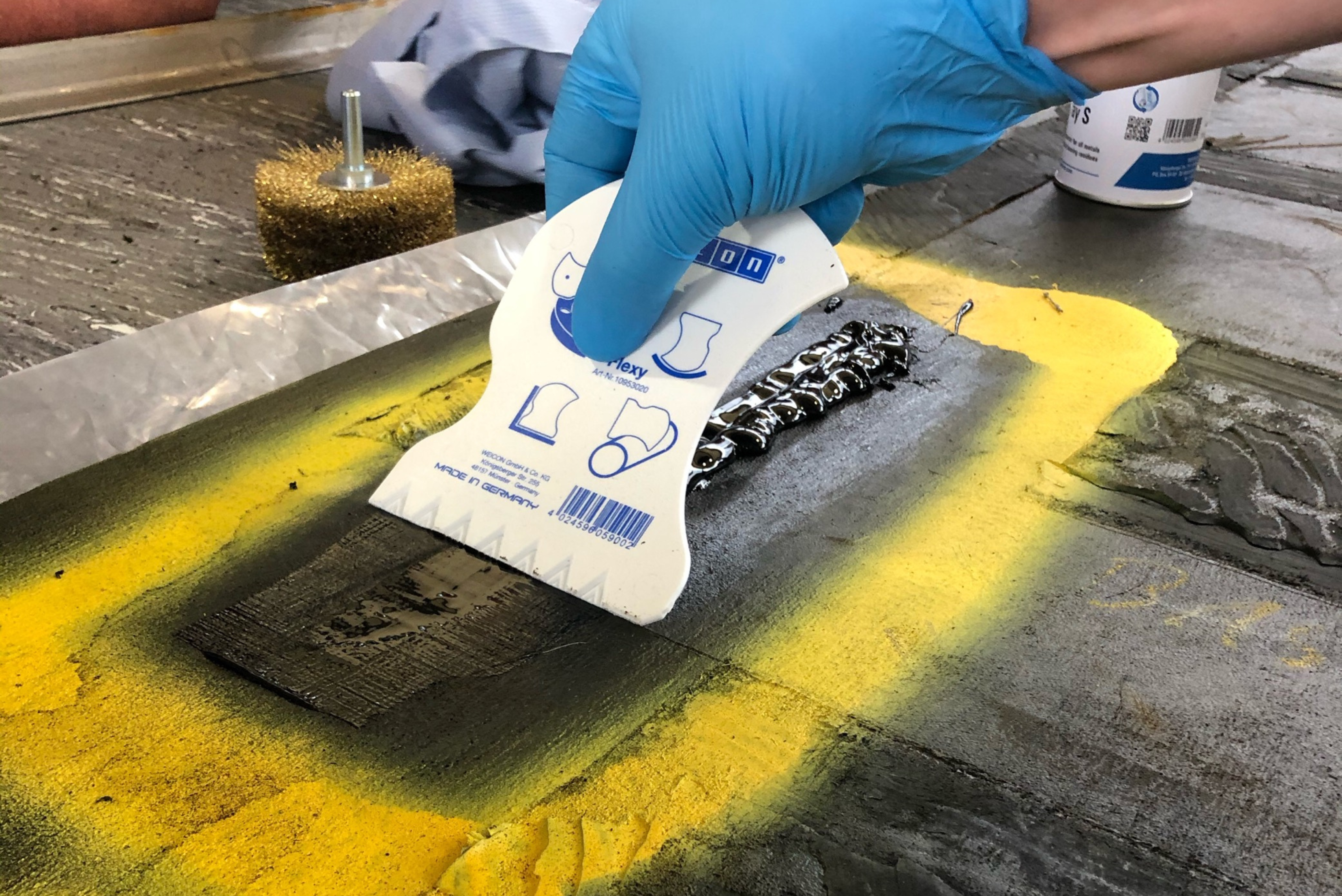 Une personne portant un gant bleu utilise une spatule en plastique blanche pour étaler ou lisser de la résine ou du mastic noir sur une surface texturée marquée de jaune.