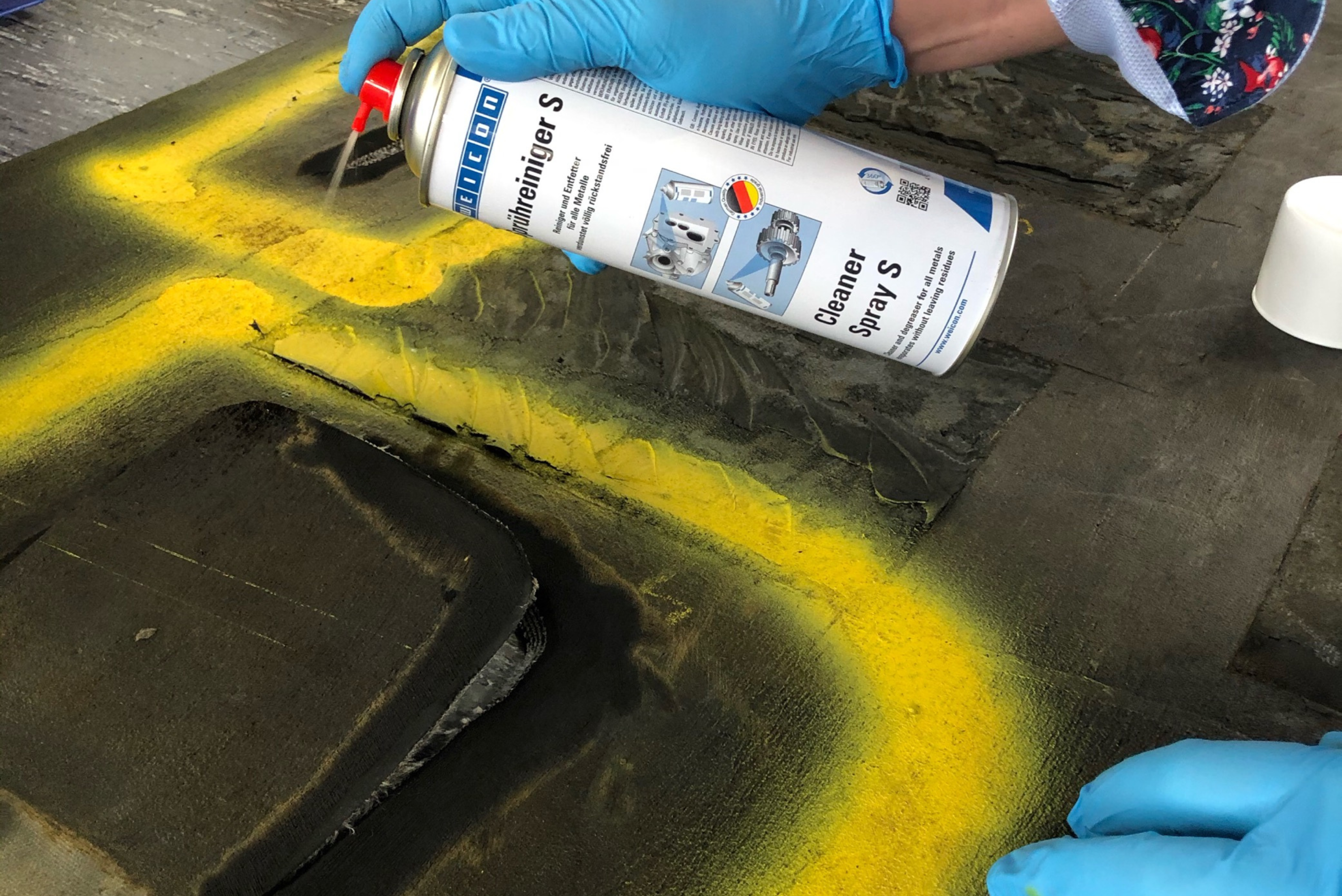 Une main portant un gant bleu utilise un aérosol de nettoyant "Cleaner Spray S" pour pulvériser un produit sur une surface sombre présentant des marquages jaunes.