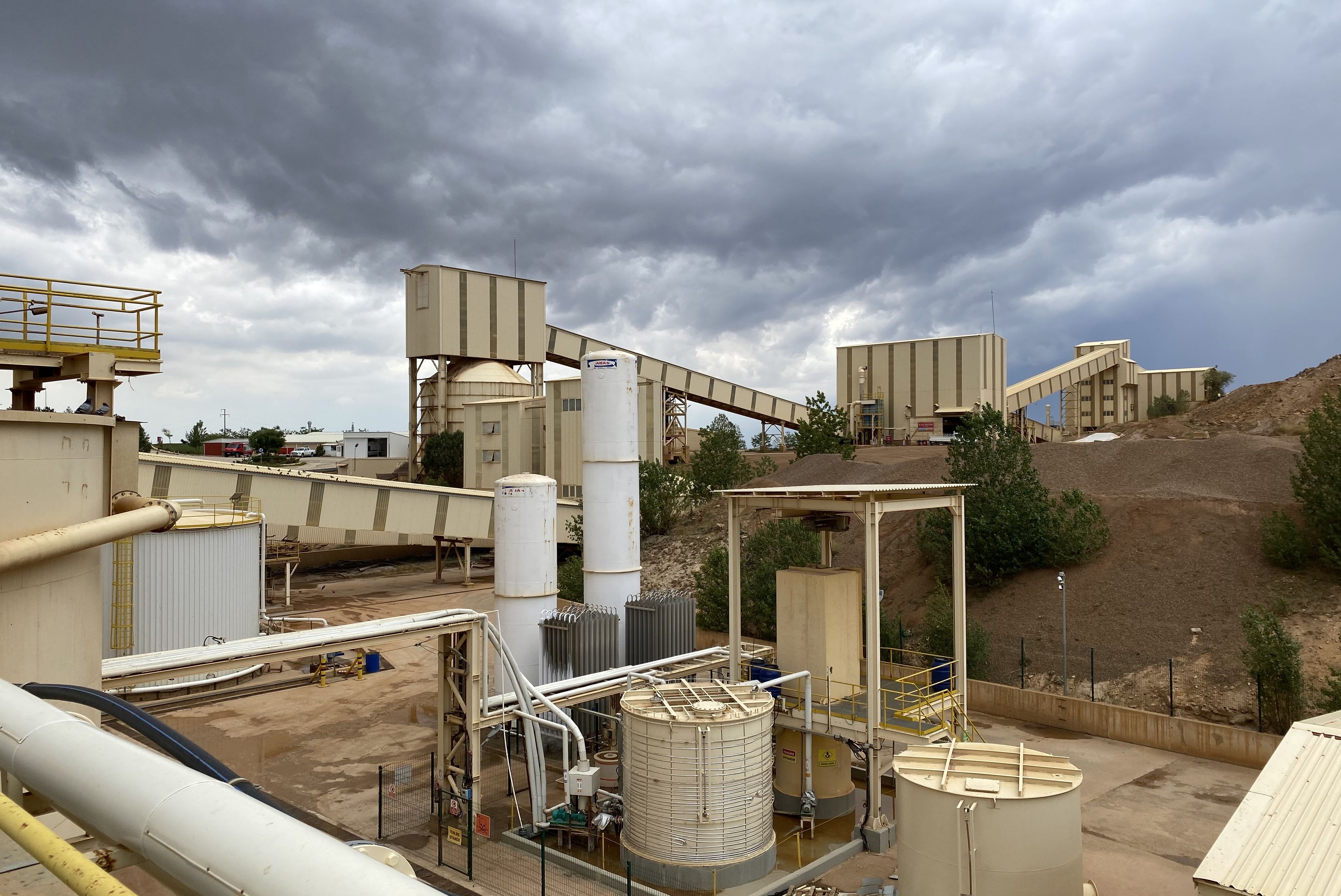 Une vue d'ensemble extérieure d'une grande installation industrielle ou d'une mine, montrant des bâtiments de traitement, des réservoirs verticaux de stockage et des bandes transporteuses surélevées sous un ciel nuageux et menaçant