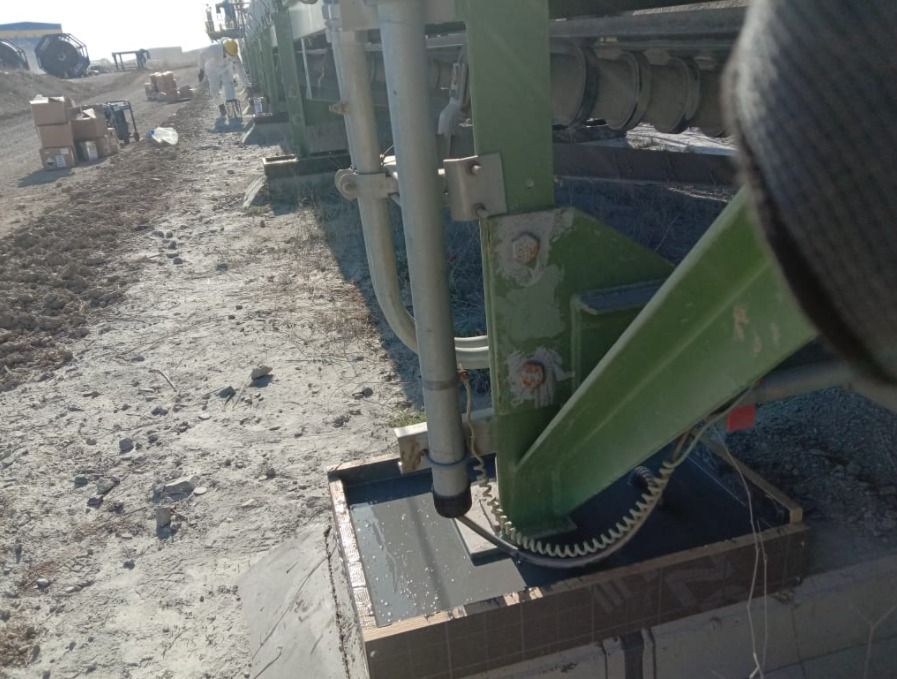 Vue en contre-plongée d'une structure de support métallique verte extérieure, avec une base solidement ancrée dans un coffrage en béton frais et des équipements industriels visibles en arrière-plan.