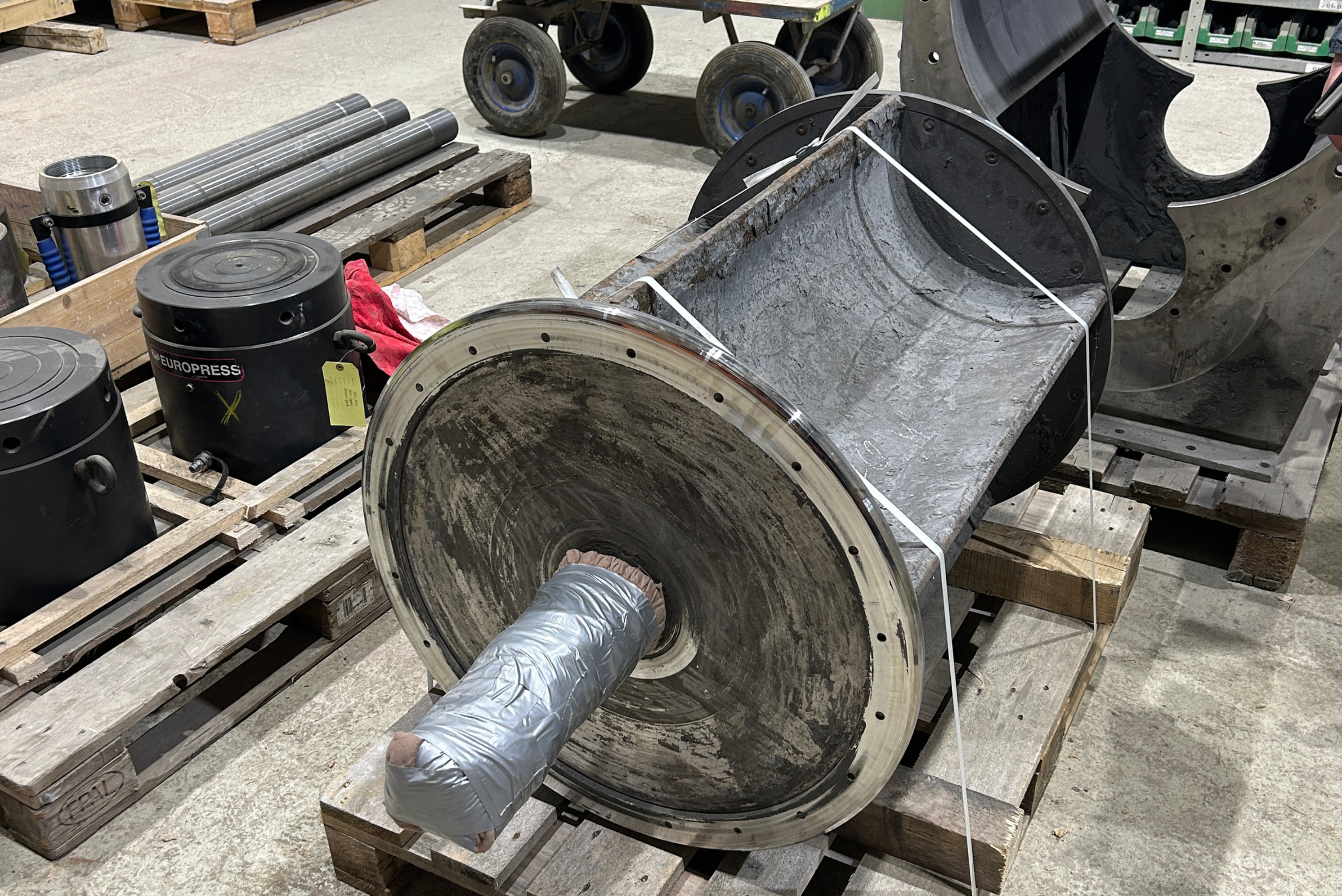 Vue rapprochée d'une grande pièce industrielle cylindrique montée sur des palettes en bois dans un atelier, dotée d'une bride circulaire percée de trous sur le côté et d'un arbre d'entraînement enveloppé de ruban adhésif gris.
